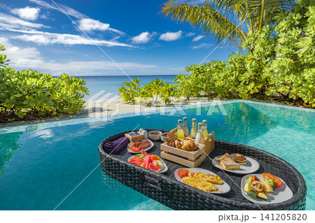 Floating breakfast basket with fresh food and drinks in private pool by the beach. Tropical morning meal on water, lush plants and turquoise sea behind. Trendy luxury vacation experience for resort Floating breakfast basket with fresh food and drinks in private pool by the beach. Tropical morning meal on water, lush plants and turquoise sea behind. Trendy luxury vacation experience for resort 141205820