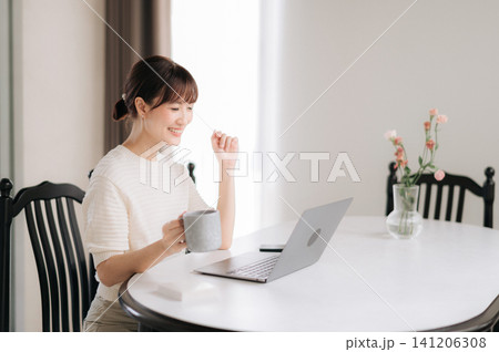 Happy asian woman enjoying modern lifestyle working on laptop computer drinking coffee from mug sitting at white table smiling with cheerful expression in bright room 141206308