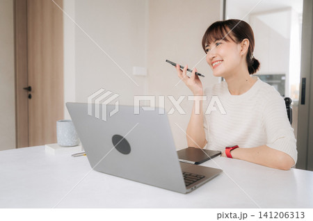 Young happy asian woman enjoying modern lifestyle working from home office smiling while talking on mobile phone sitting at bright desk with laptop computer having cheerful conversation 141206313