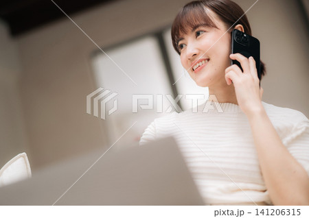 Happy young asian woman talking on mobile phone smiling working with laptop enjoying modern lifestyle sitting at desk indoor communication happiness business casual routine Happy young asian woman talking on mobile phone smiling working with laptop enjoying modern lifestyle sitting at desk indoor communication happiness business casual routine 141206315