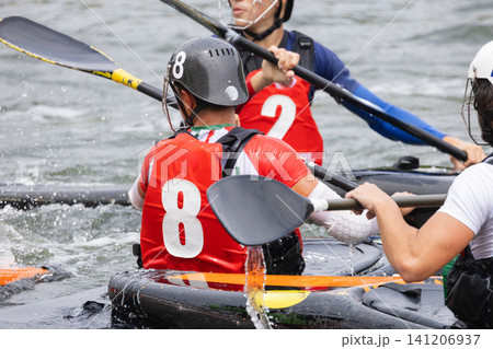 Canoe polo players in action on water, featuring athletes in red and blue jerseys, paddling with oars, and engaging in competitive gameplay during a match 141206937