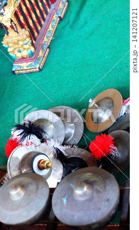 Balinese gamelan bronze cymbals, knobbed gongs, decorative tassels on green mat beside carved painted wooden instrument, traditional ceremonial music setup with rich metal detail 141207121