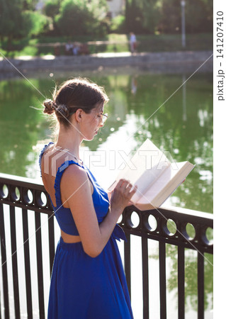 Young woman reading book in summer park. Digital detox in action. Intentional escape from screens. Brain reset through nature and words. Mindfulness among green trees. Book lovers day Young woman reading book in summer park. Digital detox in action. Intentional escape from screens. Brain reset through nature and words. Mindfulness among green trees. Book lovers day 141207410