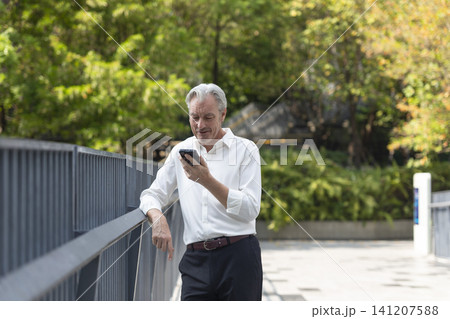 Senior adult professional business man holding smartphone, talking business and standing outdoor in a park among office buildings. Senior adult professional business man holding smartphone, talking business and standing outdoor in a park among office buildings. 141207588