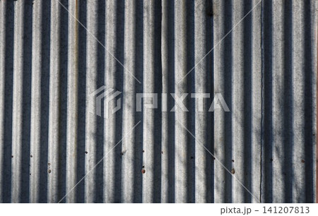 The texture of old corrugated metal sheets with vertical relief. The galvanized gray surface is covered with small spots, traces of fasteners, and soft shadows from trees. Textured metal background The texture of old corrugated metal sheets with vertical relief. The galvanized gray surface is covered with small spots, traces of fasteners, and soft shadows from trees. Textured metal background 141207813