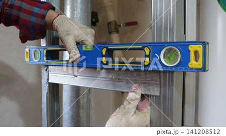 A worker checking the accuracy of the installation of galvanized metal profiles using a blue spirit level. The frame of the future wall is visible in the frame against the backdrop of utility lines. 141208512