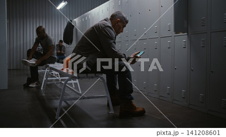 Factory Worker Sitting on Bench and Scrolling Social Media on Smartphone in Locker Room Factory Worker Sitting on Bench and Scrolling Social Media on Smartphone in Locker Room 141208601