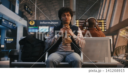 Young Male Traveler Eating Pastry and Waiting for Flight in Airport Lounge 141208915
