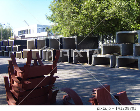 A construction site storage area features rows of rectangular precast concrete blocks alongside stacks of curved, rust-colored metal industrial components, all neatly arranged on asphalt under trees A construction site storage area features rows of rectangular precast concrete blocks alongside stacks of curved, rust-colored metal industrial components, all neatly arranged on asphalt under trees 141209703