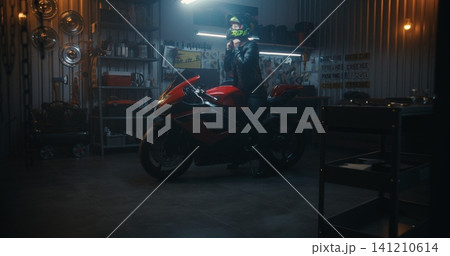 Female Biker in Leather Gear Putting on Helmet and Starting Red Sport Bike in Garage Female Biker in Leather Gear Putting on Helmet and Starting Red Sport Bike in Garage 141210614