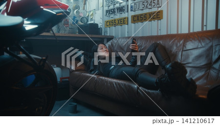 Female Lies Comfortably on Leather Couch Near Motorcycle, Enjoys Leisure Time in Garage Female Lies Comfortably on Leather Couch Near Motorcycle, Enjoys Leisure Time in Garage 141210617