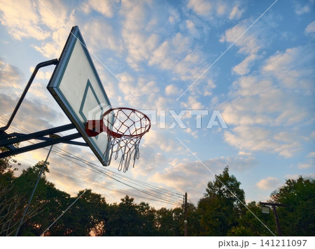 A low angle view of an outdoor basketball hoop under a dramatic evening sky with golden clouds. Perfect for sports, inspiration, motivation, and healthy lifestyle concepts. A low angle view of an outdoor basketball hoop under a dramatic evening sky with golden clouds. Perfect for sports, inspiration, motivation, and healthy lifestyle concepts. 141211097