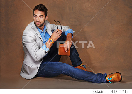 Style, book and man with glasses in studio with trendy, stylish and classy suit on floor. Reading, story and handsome male person with elegant fashion for outfit and novel by brown background. 141211712
