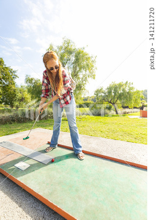 Girl plays mini golf in a sunny park while wearing sunglasses on a clear day in the afternoon during a fun outing with friends 141211720