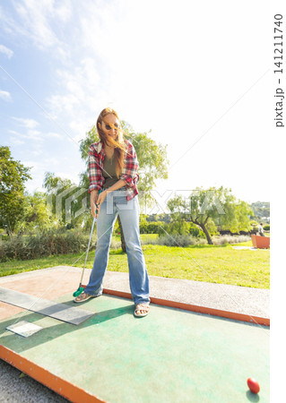 Young woman plays mini golf on a sunny day at an outdoor course surrounded by greenery and a blue sky with clouds Young woman plays mini golf on a sunny day at an outdoor course surrounded by greenery and a blue sky with clouds 141211740
