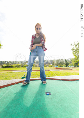 Person plays mini golf on a sunny day in an outdoor park with green grass and trees in the background 141211790