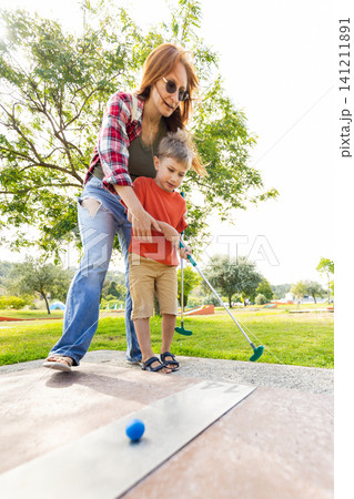 Mother and son play mini golf together at a park on a sunny day while practicing their swing and enjoying the outdoors Mother and son play mini golf together at a park on a sunny day while practicing their swing and enjoying the outdoors 141211891