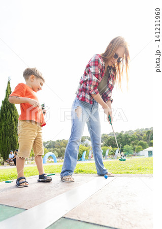 Woman and boy play mini golf together at a park on a sunny day with green trees in the background Woman and boy play mini golf together at a park on a sunny day with green trees in the background 141211960