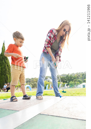 Child and adult play mini golf in a park during the afternoon with bright sunlight and green grass in the background 141211966