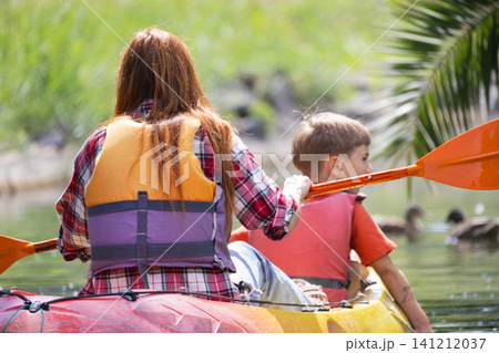 Woman and child paddle a kayak on a lake among ducks during a sunny day in a nature setting Woman and child paddle a kayak on a lake among ducks during a sunny day in a nature setting 141212037