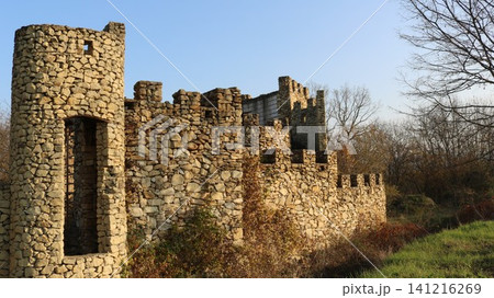 The abandoned ruins of a medieval stone castle with a round tower and crenellated walls. Sunlight illuminates the textured stonework against a backdrop of autumn trees and a clear sky. The abandoned ruins of a medieval stone castle with a round tower and crenellated walls. Sunlight illuminates the textured stonework against a backdrop of autumn trees and a clear sky. 141216269