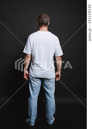 A rear view of a casually dressed man standing against a dark A rear view of a casually dressed man standing against a dark 141216536