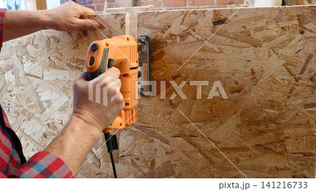 The hands of a carpenter in a plaid shirt, cutting OSB with an orange jigsaw. The craftsman confidently guides the tool along the marked line, creating a straight vertical cut. 141216733
