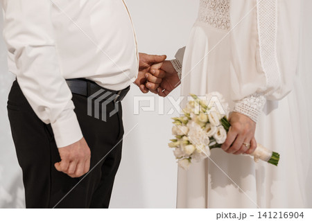 Groom and Bride Holding Hands with White Rose Bouquet in Minimalist Studio Setting 141216904