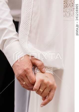 Close up of a couple holding hands showing gold wedding rings and white clothing in a soft romantic style. Close up of a couple holding hands showing gold wedding rings and white clothing in a soft romantic style. 141216905