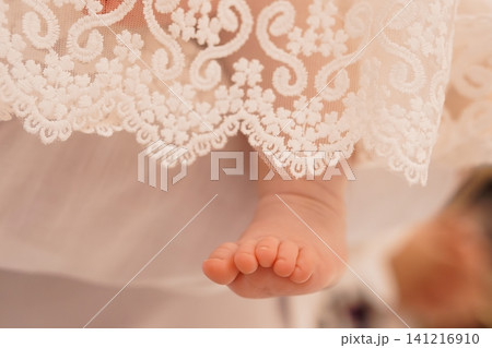 Close up of newborn baby foot peaking from white lace baptismal gown in soft warm lighting. Close up of newborn baby foot peaking from white lace baptismal gown in soft warm lighting. 141216910