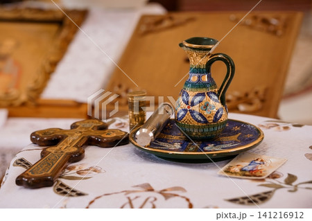Orthodox Christian ritual objects including an ornate ceramic pitcher and wooden cross on a white altar cloth. Orthodox Christian ritual objects including an ornate ceramic pitcher and wooden cross on a white altar cloth. 141216912