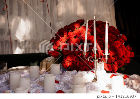 Romantic arrangement of red roses bouquet and lit white candles on a white fabric table with silver candelabra. Romantic arrangement of red roses bouquet and lit white candles on a white fabric table with silver candelabra. 141216937