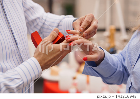 Man putting diamond engagement ring on womans finger from red jewelry box outdoors in bright daylight. Man putting diamond engagement ring on womans finger from red jewelry box outdoors in bright daylight. 141216948