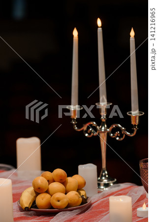 Lit white taper candles in silver candelabra with fresh yellow fruit on red tablecloth against dark background. Lit white taper candles in silver candelabra with fresh yellow fruit on red tablecloth against dark background. 141216965
