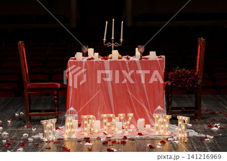 Romantic marriage proposal dinner table with glowing letters and red roses on a dark theater stage. 141216969