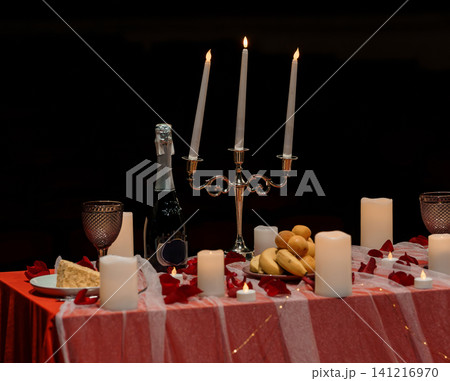 Romantic dinner table with champagne bottle, lit candles and red rose petals on red linen background. Romantic dinner table with champagne bottle, lit candles and red rose petals on red linen background. 141216970