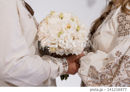 Couple in traditional embroidered white linen shirts holding a white flower bouquet against a plain background. Couple in traditional embroidered white linen shirts holding a white flower bouquet against a plain background. 141216977