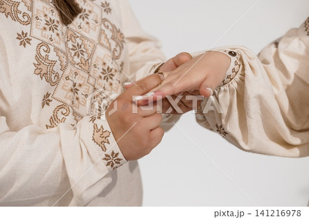 Person placing a ring on a finger wearing traditional embroidered linen clothing against a white background. Person placing a ring on a finger wearing traditional embroidered linen clothing against a white background. 141216978
