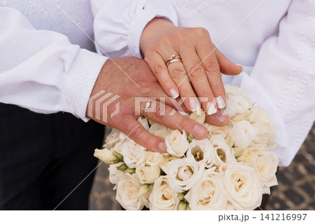 Close up of couple hands with wedding rings holding a white rose bouquet in traditional white clothing. 141216997
