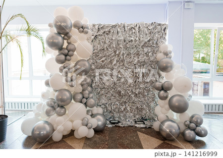Modern silver and white balloon arch framing a shimmering metallic tinsel backdrop in a bright indoor space. Modern silver and white balloon arch framing a shimmering metallic tinsel backdrop in a bright indoor space. 141216999