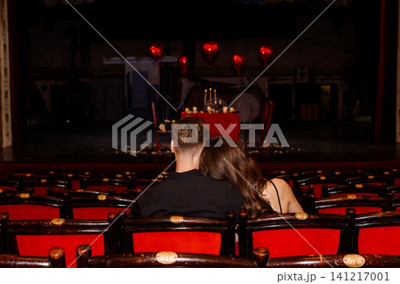 Romantic couple sitting in red theater seats facing a dinner table with heart balloons on a dark stage. 141217001