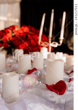 Romantic arrangement of white candles and red rose petals on white satin fabric with a rose bouquet background. Romantic arrangement of white candles and red rose petals on white satin fabric with a rose bouquet background. 141217011