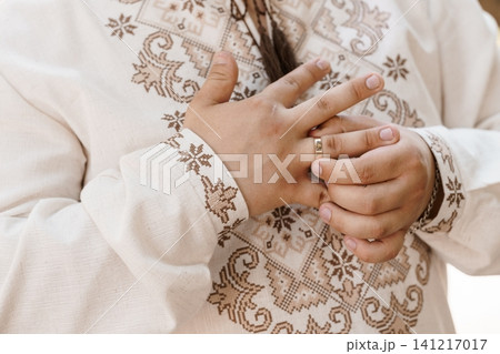 Man wearing traditional embroidered linen shirt touching gold wedding ring on finger in soft natural light. Man wearing traditional embroidered linen shirt touching gold wedding ring on finger in soft natural light. 141217017