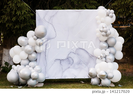 Elegant white and silver balloon arch framing a marble pattern photo backdrop outdoors in a garden. Elegant white and silver balloon arch framing a marble pattern photo backdrop outdoors in a garden. 141217020