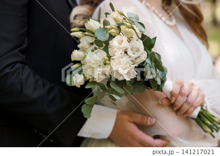 Groom in black suit embracing bride holding white floral wedding bouquet with roses and eucalyptus leaves. Groom in black suit embracing bride holding white floral wedding bouquet with roses and eucalyptus leaves. 141217021