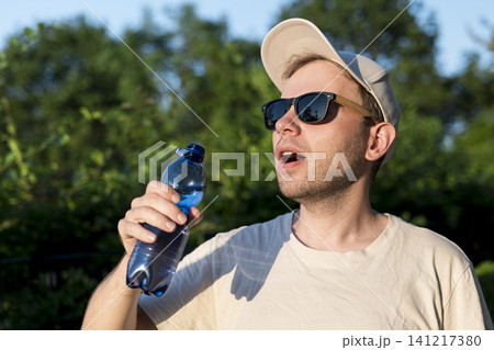 Young man wearing a cap and sunglasses drinking water from a plastic bottle outdoors, illustrating summer lifestyle adaptations. 141217380
