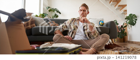 Portrait of man thinking and looking at his luggage, sitting with opened suitcase, packing for holiday Portrait of man thinking and looking at his luggage, sitting with opened suitcase, packing for holiday 141219078