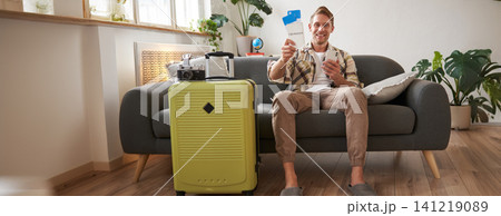 Portrait of happy man checked in online on smartphone, showing tickets in his hand, sitting near suitcase, going on vacation, preparing for holiday Portrait of happy man checked in online on smartphone, showing tickets in his hand, sitting near suitcase, going on vacation, preparing for holiday 141219089