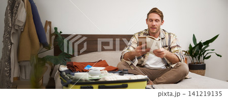 Portrait of man with suitcase, sitting and packing things for holiday, checking list of items for vacation, writing down, making notes in notebook Portrait of man with suitcase, sitting and packing things for holiday, checking list of items for vacation, writing down, making notes in notebook 141219135