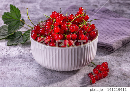 Fresh red currants in white ceramic bowl on textured gray surface with green leaves and purple cloth Fresh red currants in white ceramic bowl on textured gray surface with green leaves and purple cloth 141219411